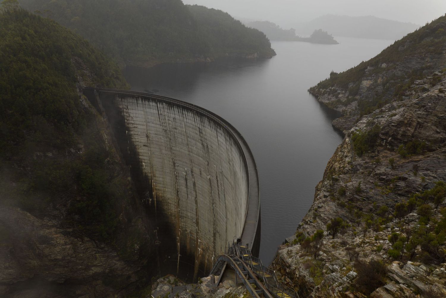 Curved concrete arch dam spanning a steep rocky gorge, holding back a calm reservoir surrounded by misty, forested hills.