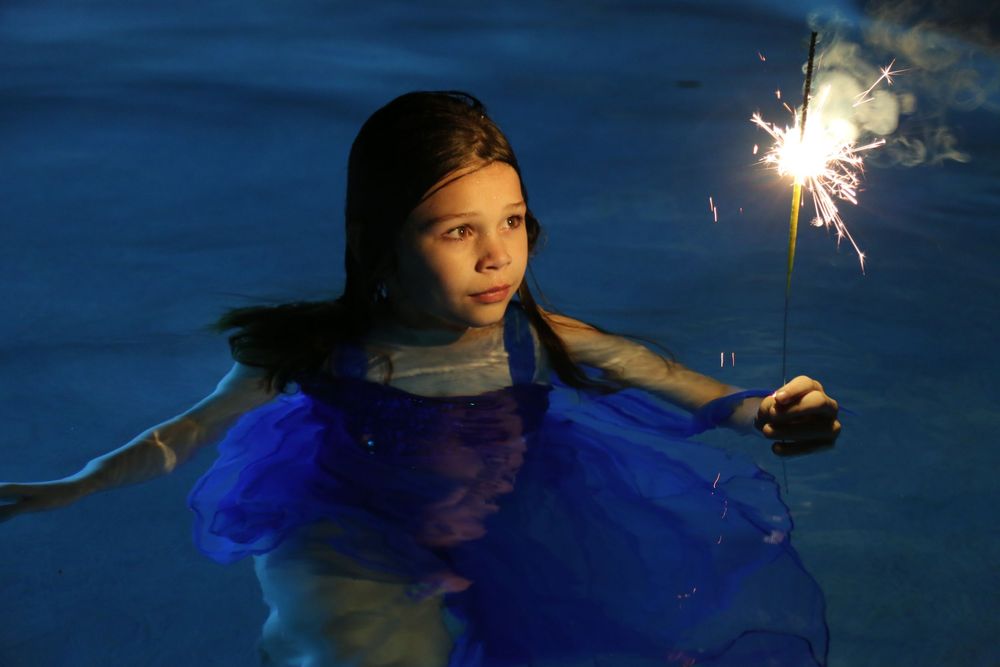 Young girl in a blue tulle dress stands in dark water at night, holding a bright sparkler that glows beside her.