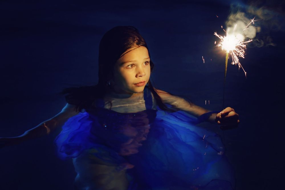 Young girl in a flowing blue dress floats in dark water at night, holding a bright sparkler that glows and sends smoke into the air.