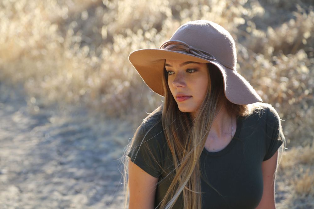 Young woman with long brown hair wearing a wide-brim mauve floppy hat and dark t-shirt, standing outdoors against golden dry grass.