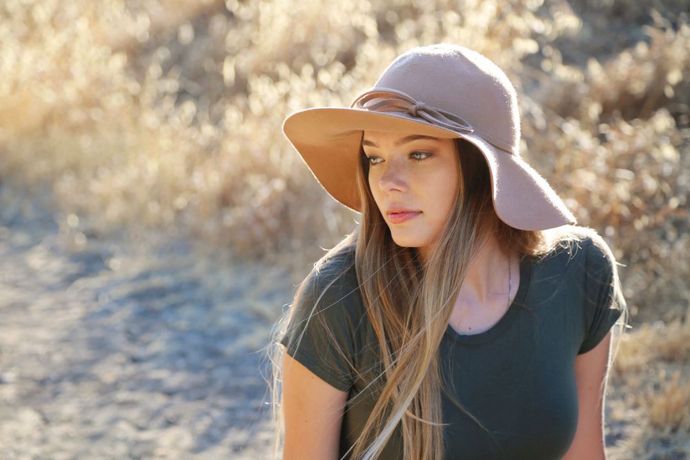 Young woman with long hair wearing a wide-brim blush hat and olive green t-shirt, standing outdoors in golden dry grass in warm sunlight.