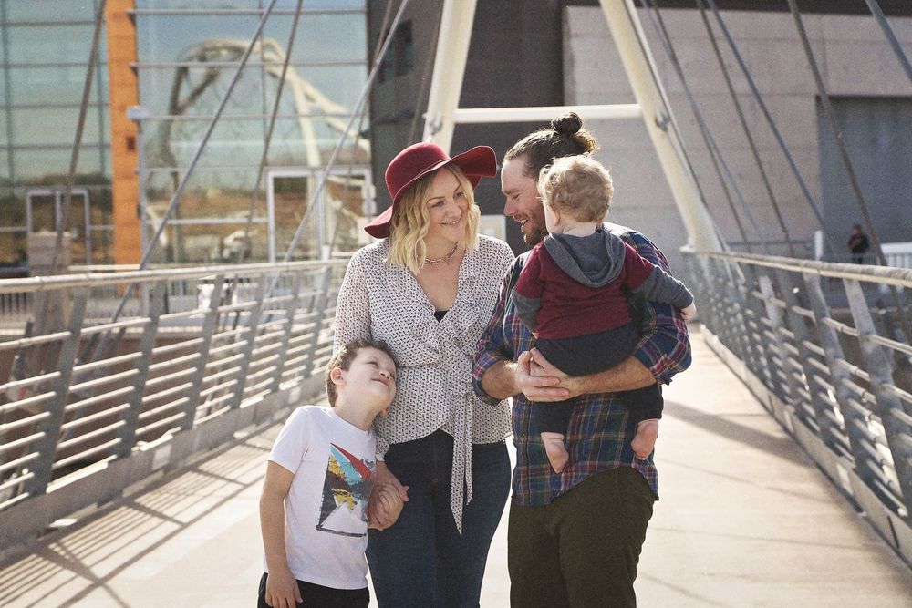 A smiling family of four walks together along a sunny pedestrian bridge, with a woman in a red hat and a man carrying a toddler.