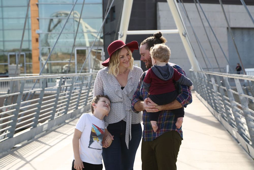 Smiling family of four walking together on a modern pedestrian bridge, with a mother in a red hat, a father carrying a toddler, and a boy.