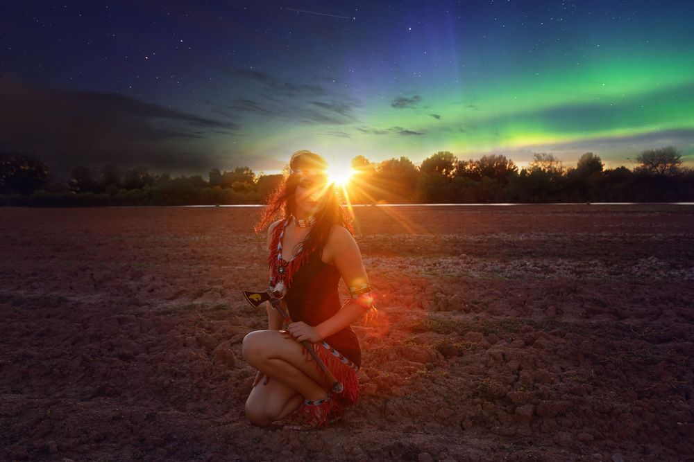 Woman in a red dress kneeling in an open field at sunset beneath a starry sky with glowing green northern lights.