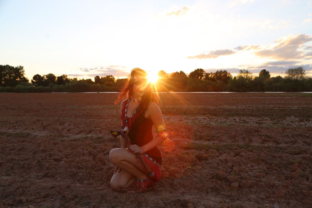 Woman in a red outfit crouching in a plowed field at sunset, with golden sunlight glowing behind distant trees.