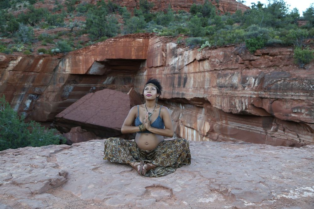 Woman meditating cross-legged in a bikini top and patterned pants on a flat red rock ledge beside sandstone cliffs and green desert trees.
