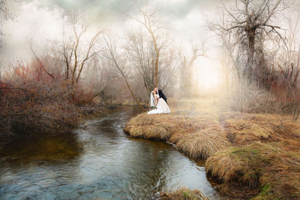 Bride in a white dress kneeling beside a calm stream in a misty autumn landscape with bare trees and golden grass at sunrise.