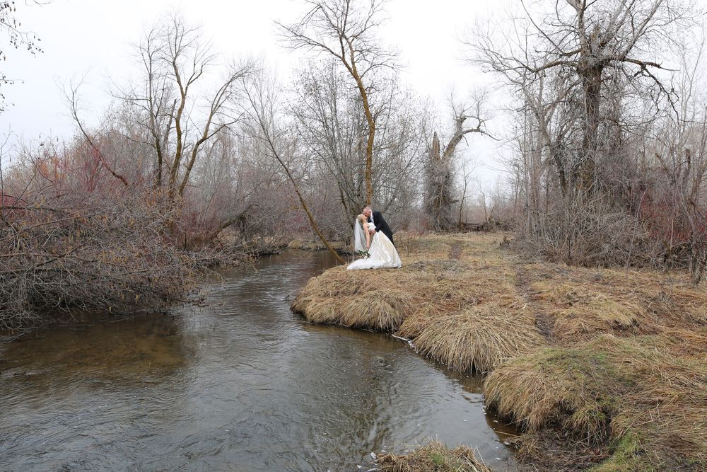 Bride in a white gown and groom in dark attire embrace beside a winding creek in a late autumn landscape with bare trees.