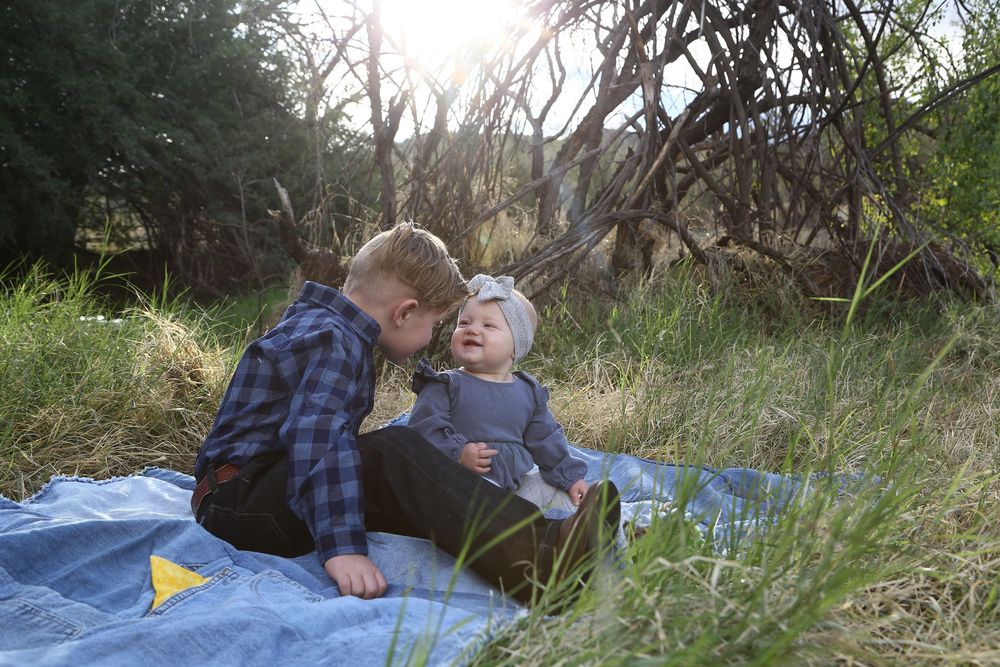 Young boy in a plaid shirt sits on a blue blanket in tall grass, smiling at a baby in a gray hat outdoors.