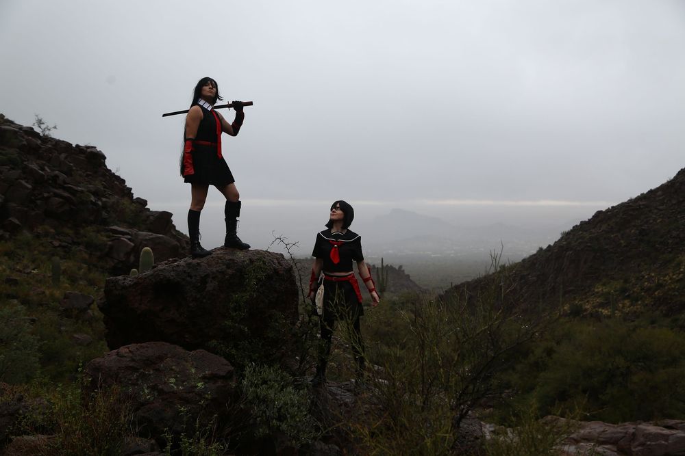 Two cosplayers in dark red costumes pose on rocky hillside terrain, one holding a staff overhead, under a moody overcast sky.