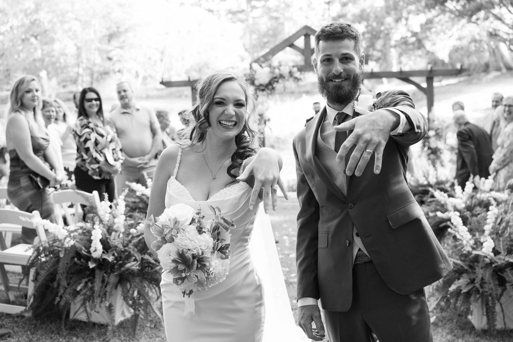 Joyful bride and groom show off their wedding rings while walking down the outdoor aisle past smiling guests and floral arrangements.