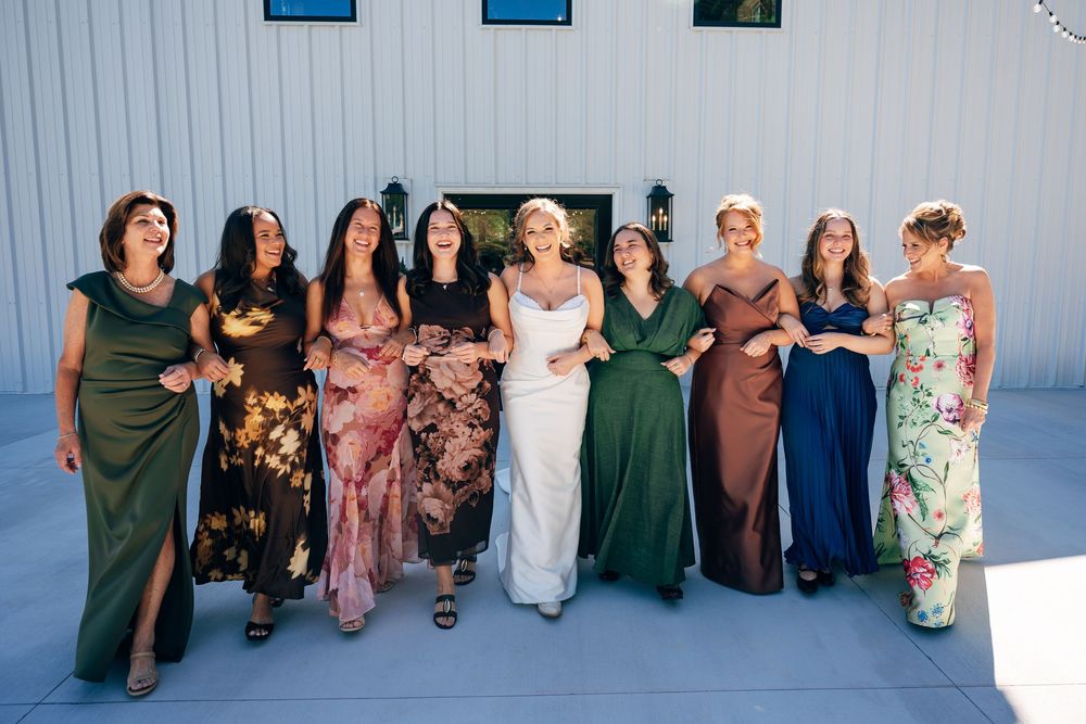 Eight smiling women in mismatched floral and solid-colored dresses stand arm in arm outdoors in front of a white building.