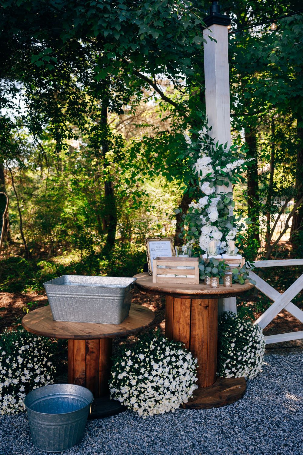 Rustic outdoor wedding decor with wooden spool tables, galvanized tubs, white flowers and a floral-wrapped post under trees.