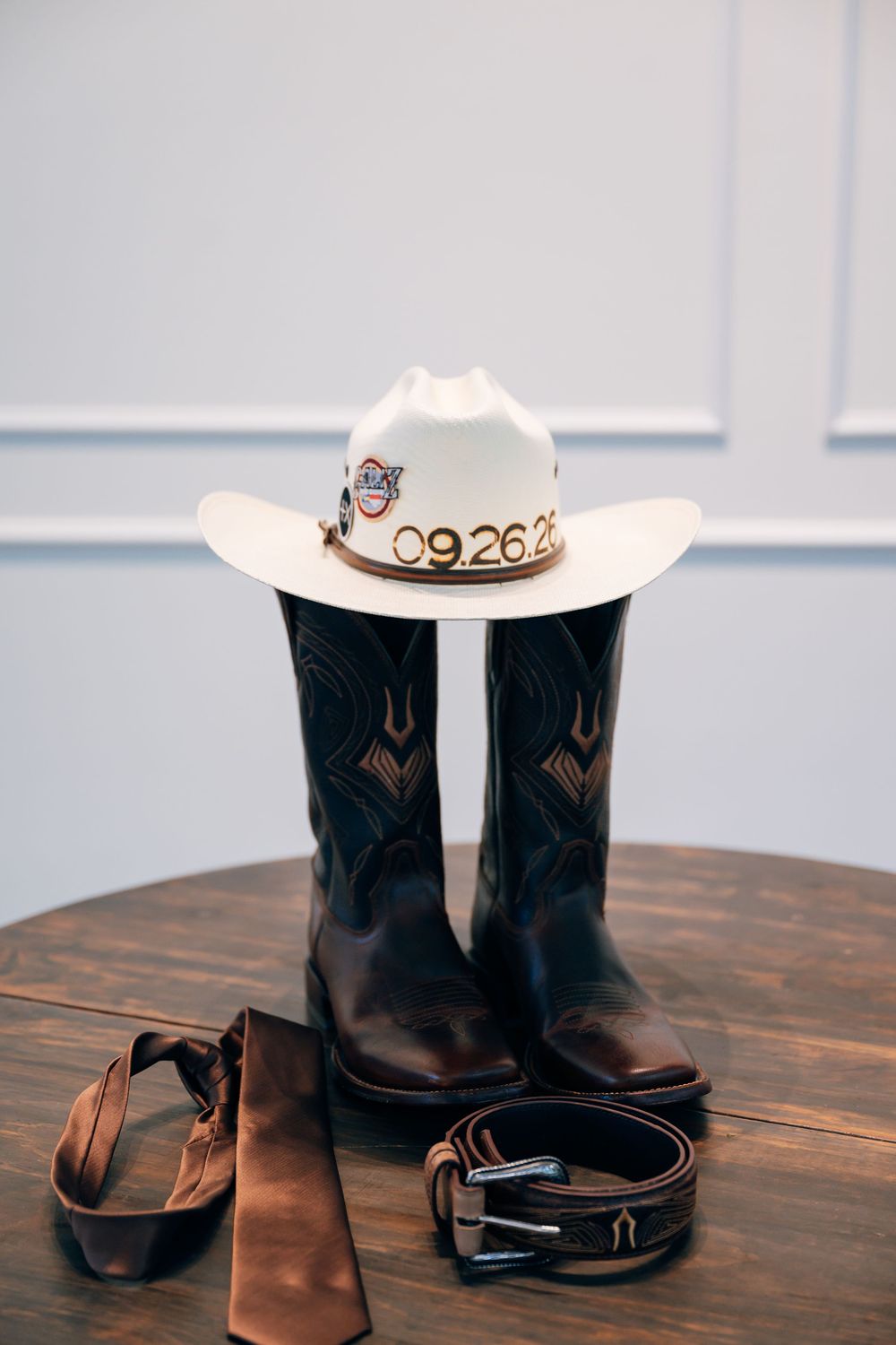 A white cowboy hat with a 09.26.21 date band rests on brown leather boots beside a satin tie and belt on a wooden table.