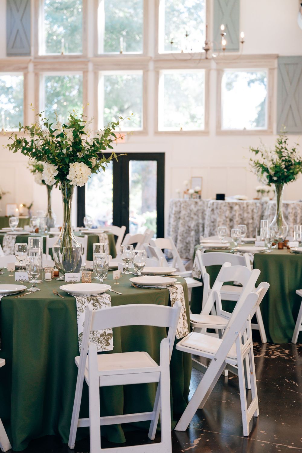 Elegant wedding reception with olive green linens, white folding chairs, and tall floral centerpieces in a bright venue with large windows.