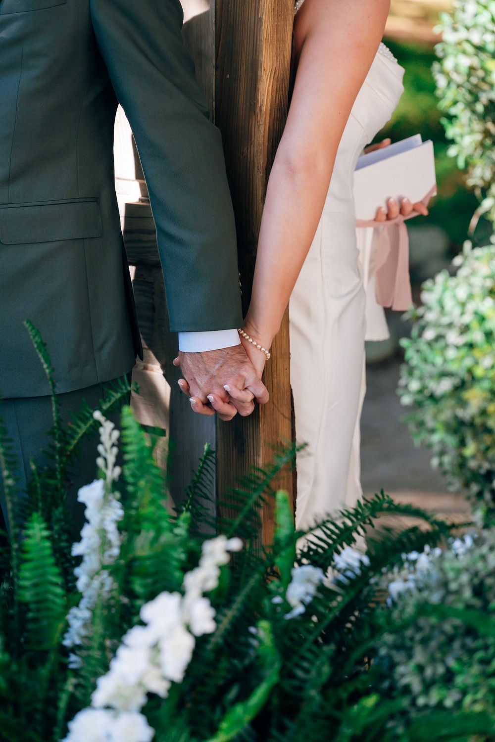 Bride and groom holding hands beside a wooden post at an outdoor wedding ceremony, with white flowers and ferns in the foreground.