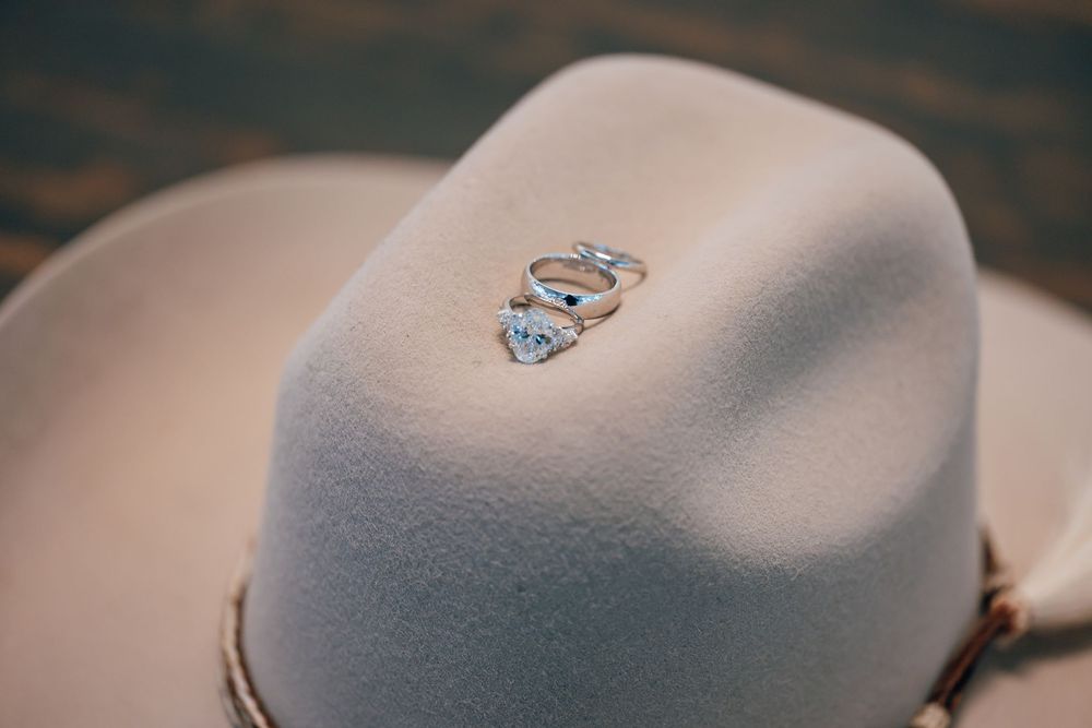 Two wedding rings, including a diamond engagement ring, rest on top of a cream felt cowboy hat with a braided leather hatband.