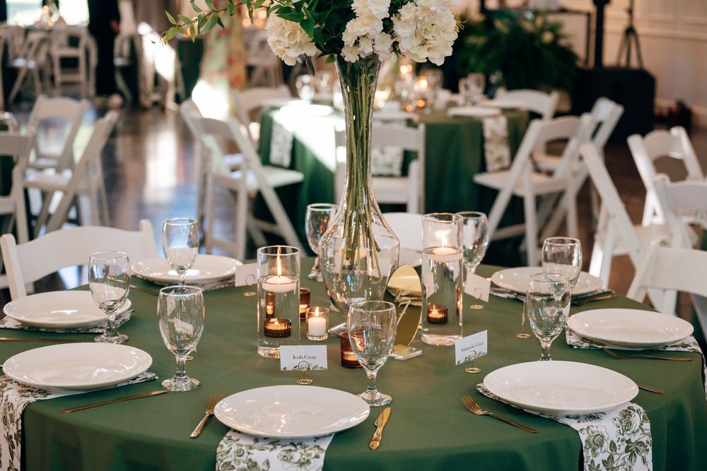 Elegant wedding reception table with emerald green linen, white plates, glassware, candles and a tall white floral centerpiece.