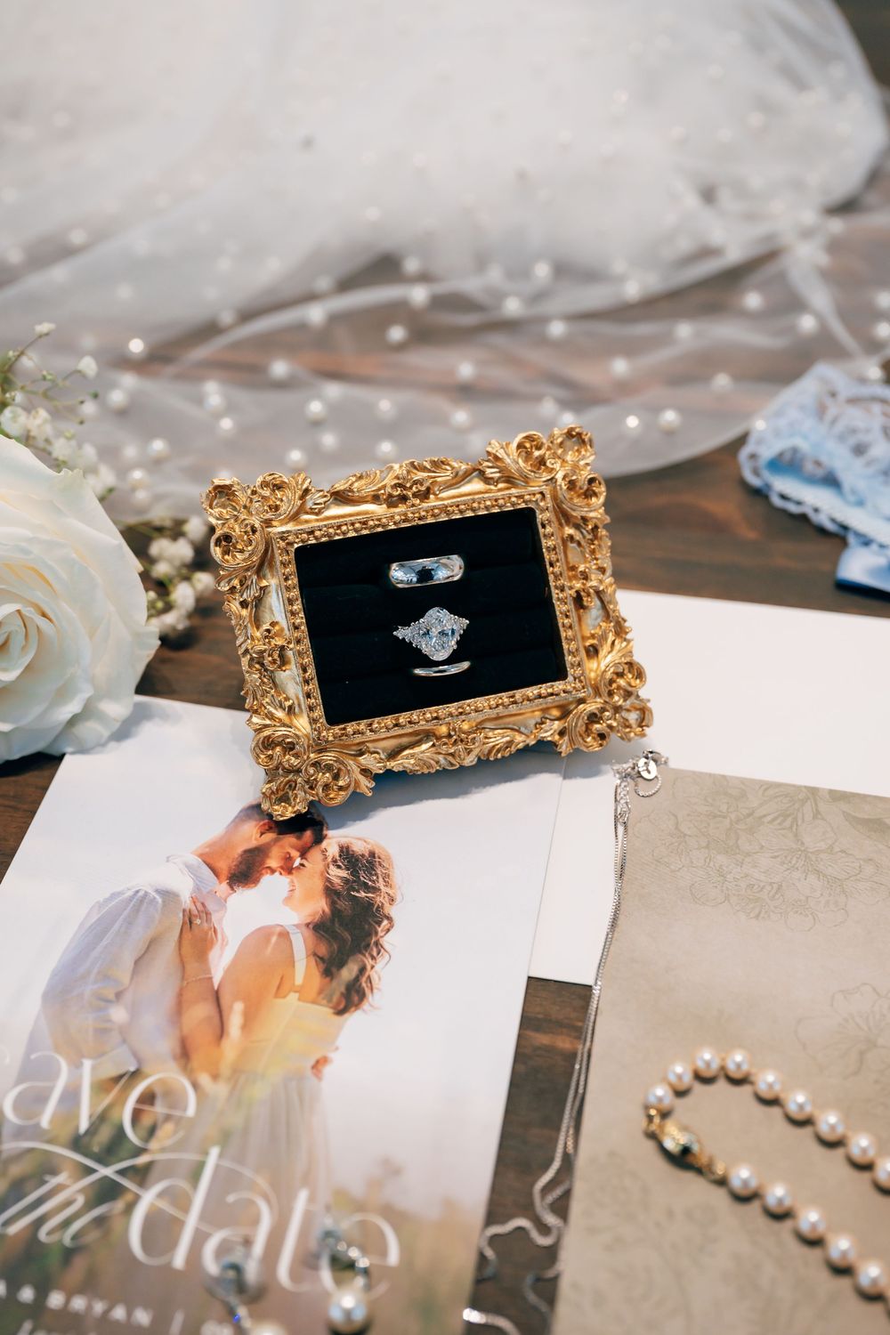 Wedding rings in a gold ornate frame ring box beside a pearl bracelet, blue garter, veil and a save the date card.