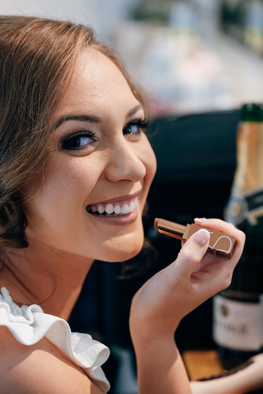Smiling bride with elegant makeup holds a lipstick tube beside a champagne bottle while getting ready for her wedding.