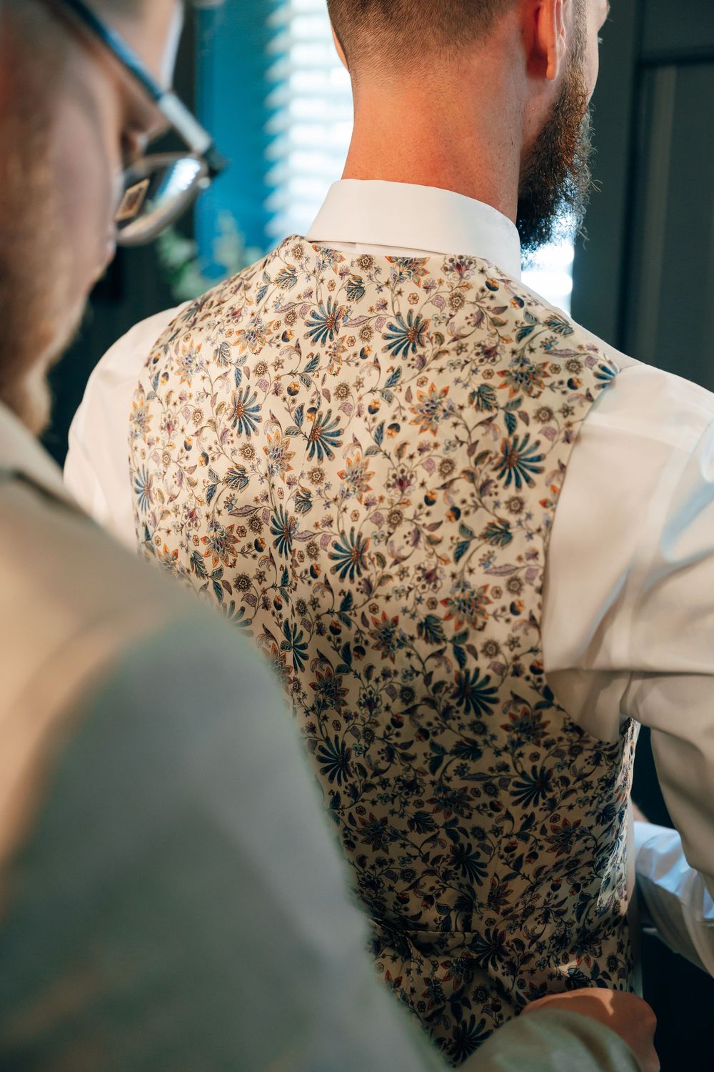 Groom in a white shirt and floral patterned vest from behind while another man in glasses helps him get ready.