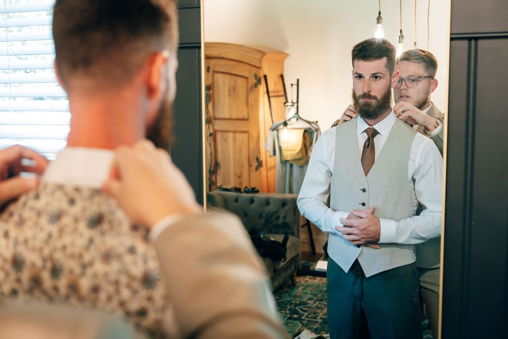 Groomsmen in vests and ties help each other get ready in front of a mirror, adjusting collars and sharing a happy moment before a wedding.