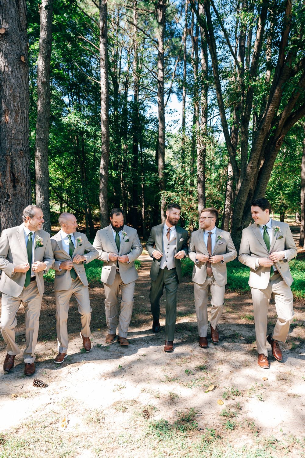 Groomsmen in tan suits and green ties laugh and walk together along a sunny forest path at an outdoor wedding.