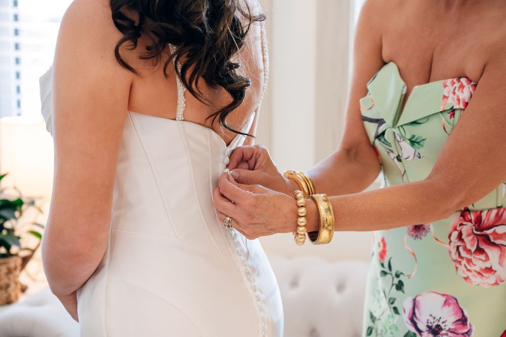 Woman in a floral mint dress wearing gold bracelets helps a bride button the back of her white wedding gown.