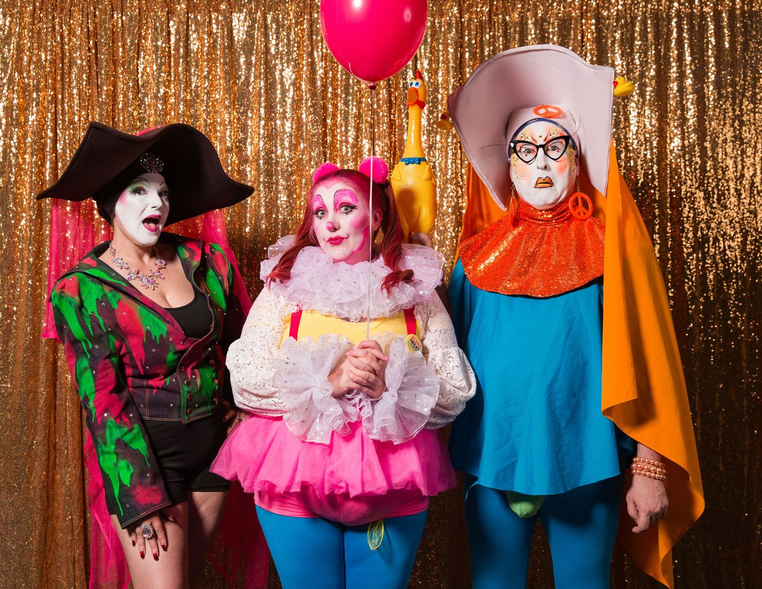 Three performers in colorful clown costumes and white face paint pose before a gold sequin backdrop, one holding a red balloon.