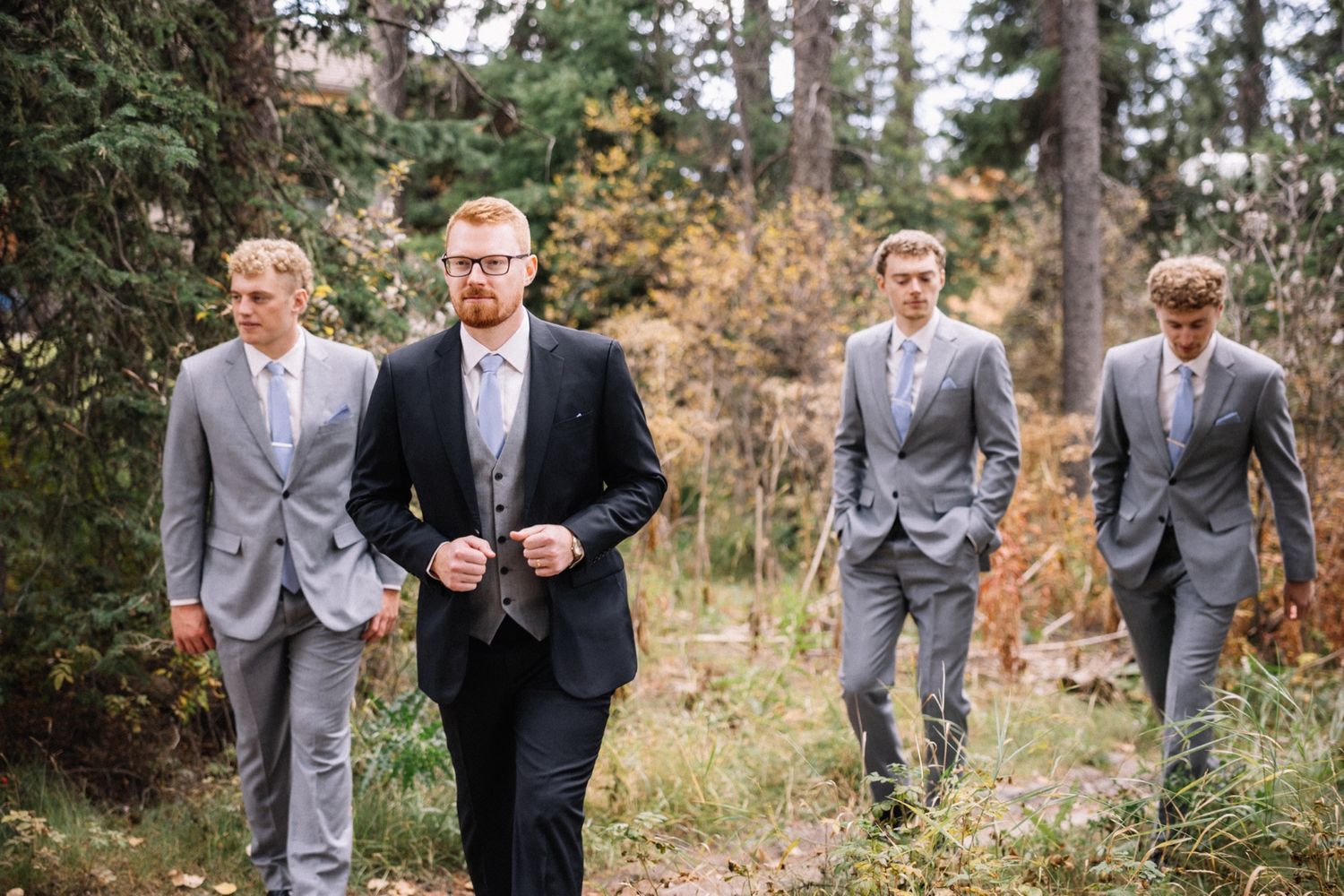 Groom and groomsmen in grey suits walking through the forest in Lake Louise