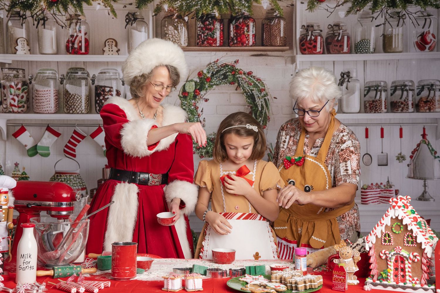 Three generations of women in Christmas outfits bake and decorate cookies together at a festive kitchen table beside a gingerbread house.