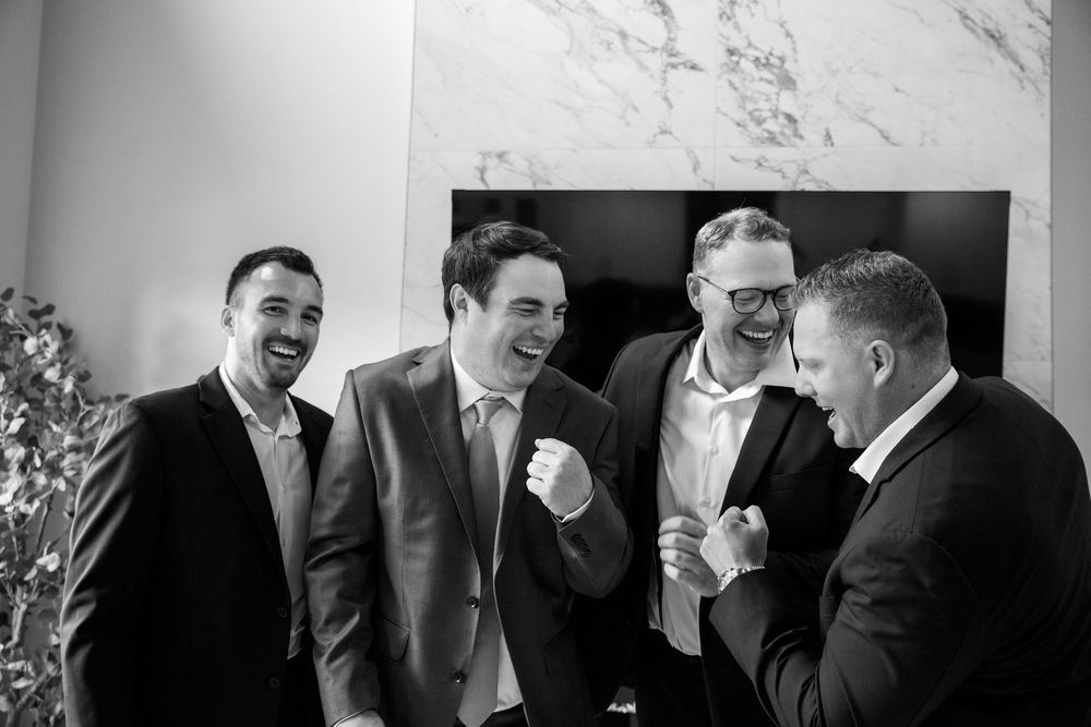Four groomsmen in dark suits laugh and celebrate together in a black and white wedding portrait.