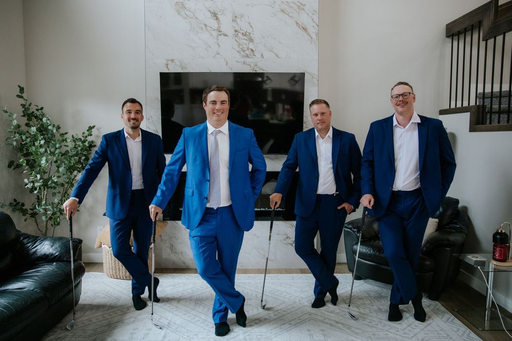 Four groomsmen in matching royal blue suits stand in a bright modern room, leaning casually on canes before the wedding.