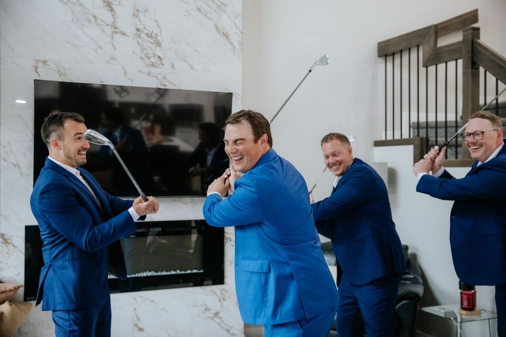 Groomsmen in blue suits laugh and swing golf clubs indoors while celebrating together before the wedding ceremony.