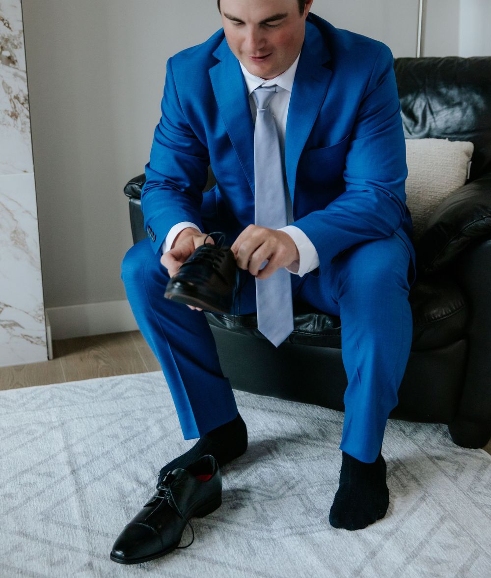 Groom in a bright blue suit and gray tie sits on a black leather chair, putting on his black dress shoes before the wedding.