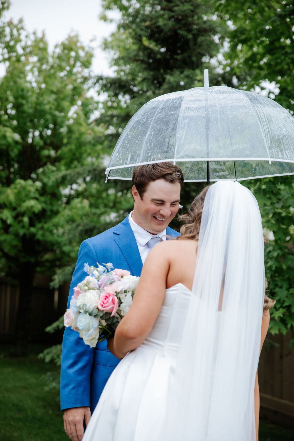 Smiling groom in a blue suit looks at his bride in a white gown and veil, holding a bouquet under a clear umbrella on a rainy wedding day.