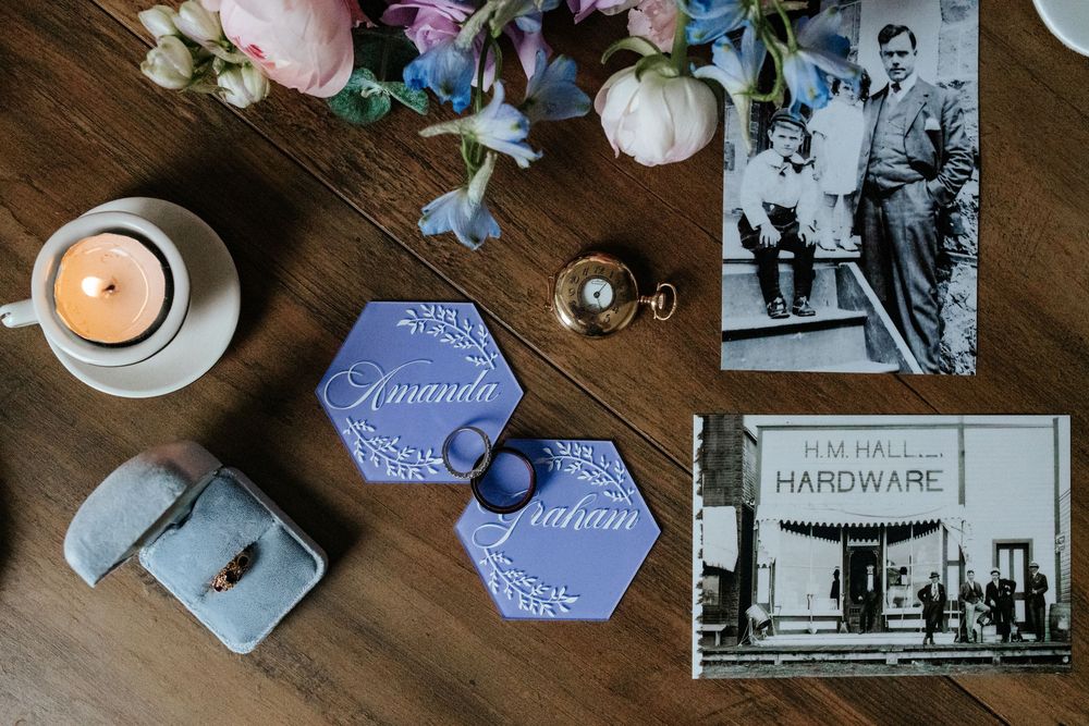 Wedding flat lay with blue hexagon name cards, a ring box, flowers, a lit candle, and vintage black-and-white photos on a wooden table.