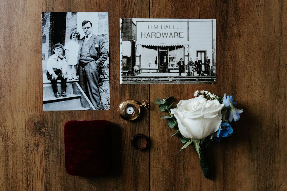 Vintage black and white photos, a gold pocket watch, rings and a white rose boutonniere styled on a rustic wood surface for a wedding.
