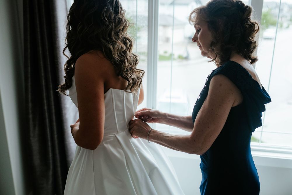 Woman in a navy dress helps a bride in a white strapless gown button up her dress beside a bright window on the wedding day.