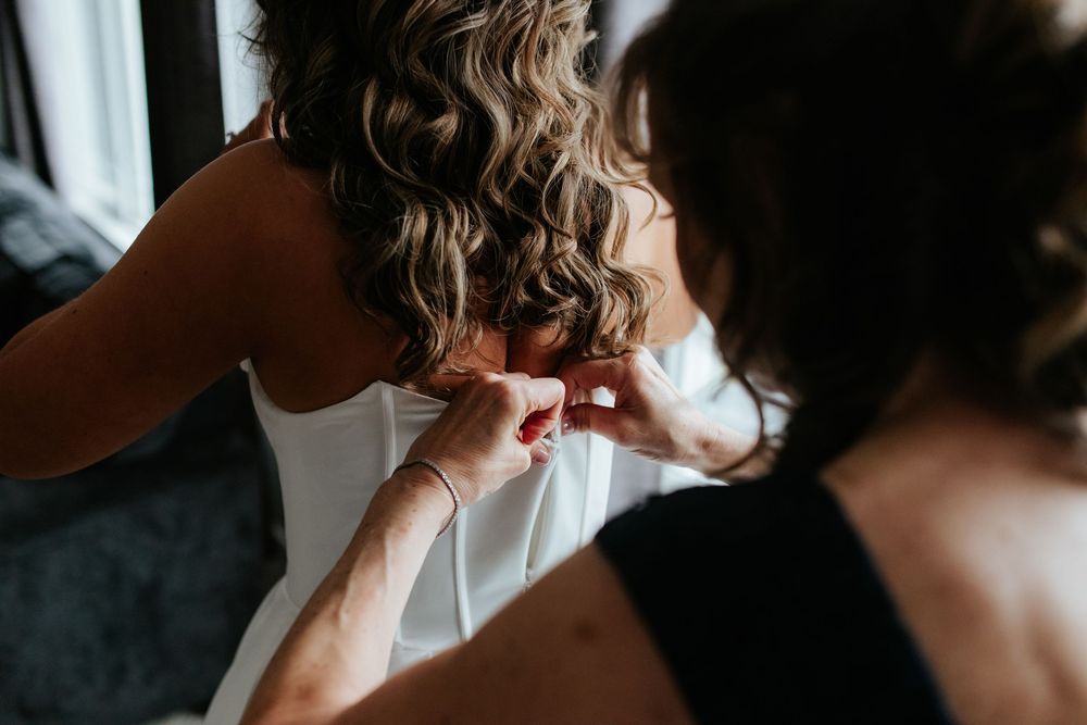 A bridesmaid helps fasten the back of the bride's white strapless wedding gown as she gets ready for the ceremony.