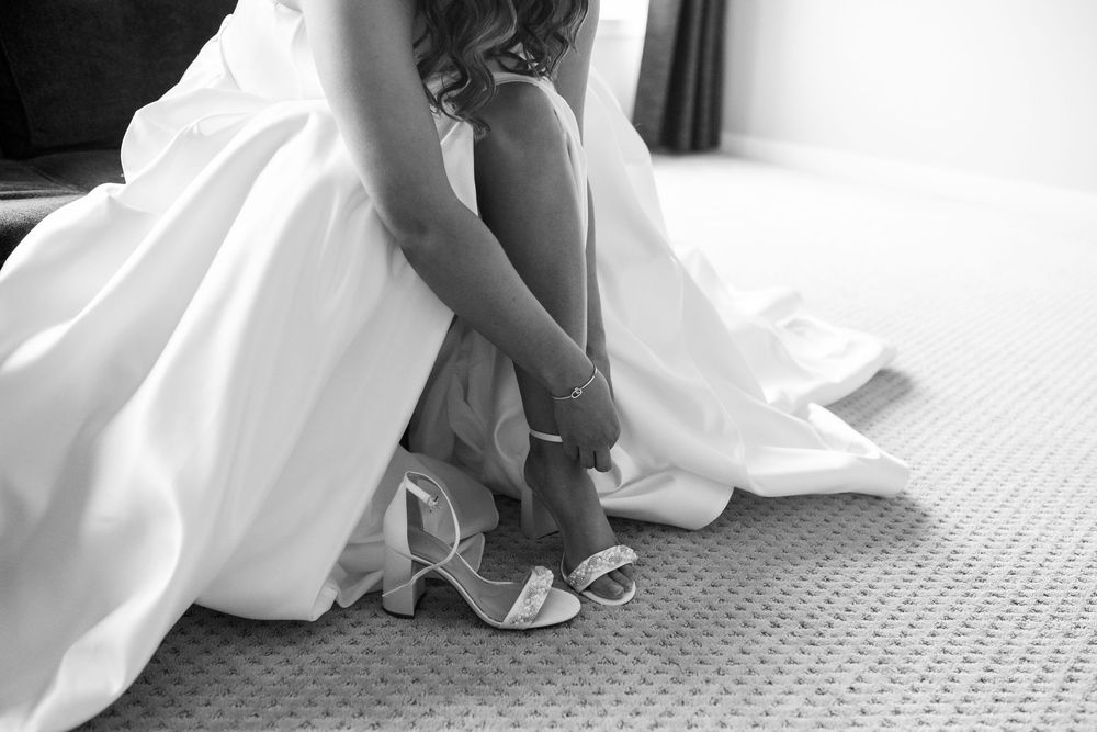 Black and white photo of a bride in a flowing satin wedding gown buckling her strappy block heel sandal on a carpeted floor.