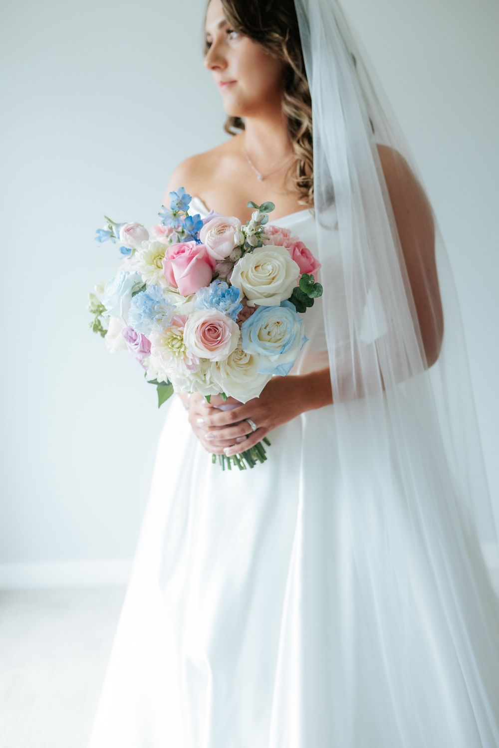Bride in a white satin gown and long veil holds a pastel bouquet of pink, blue and cream roses while gazing to the side.