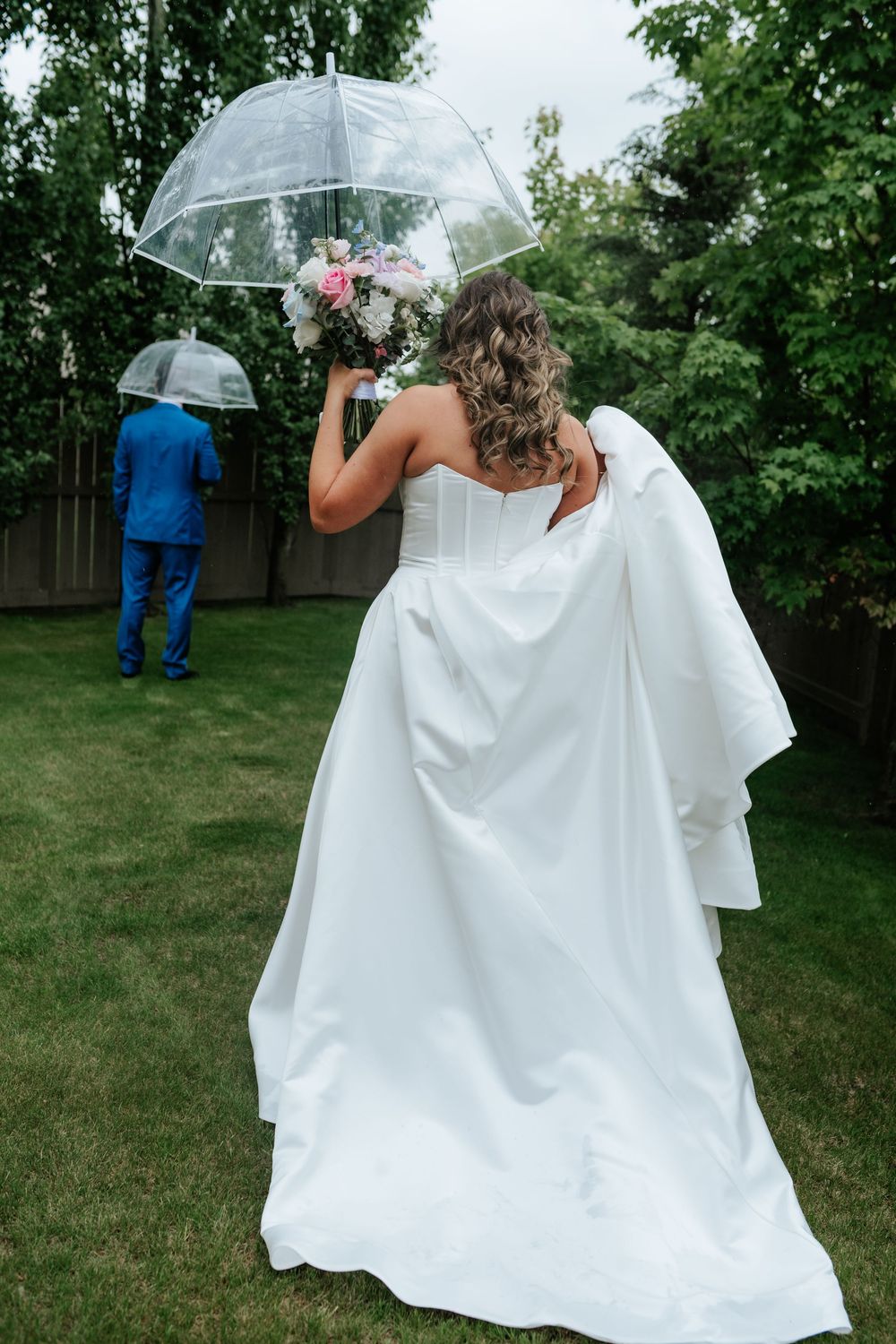 Bride in a white strapless gown holds a bouquet under a clear umbrella while the groom waits with his back turned in a garden.