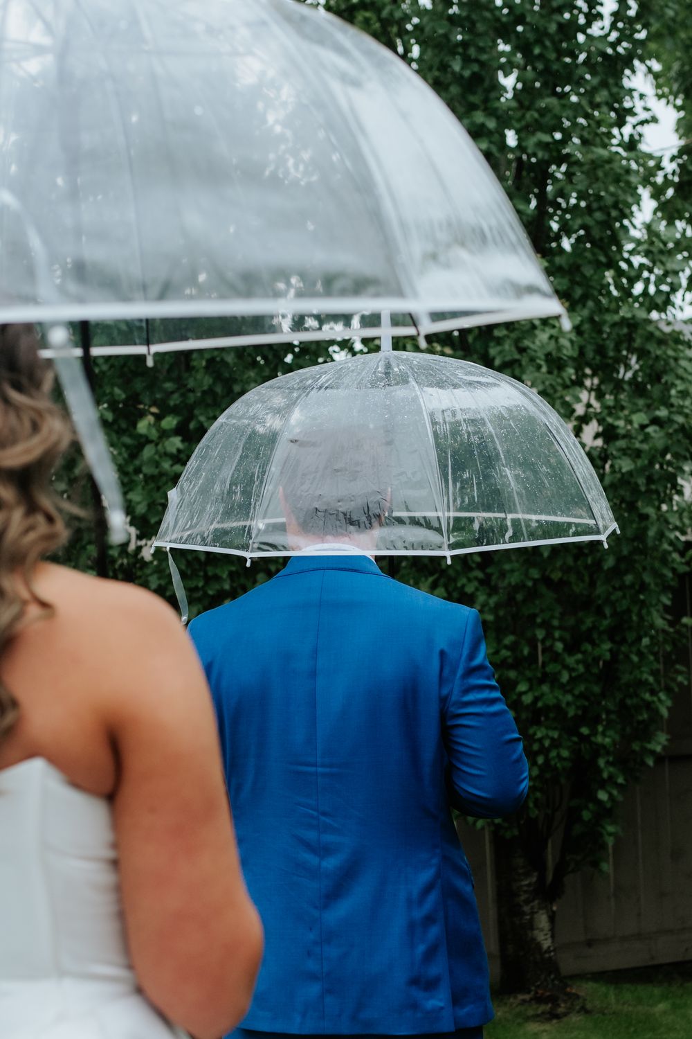 A groom in a bright blue suit walks under a clear umbrella in the rain as the bride follows behind in her white dress.