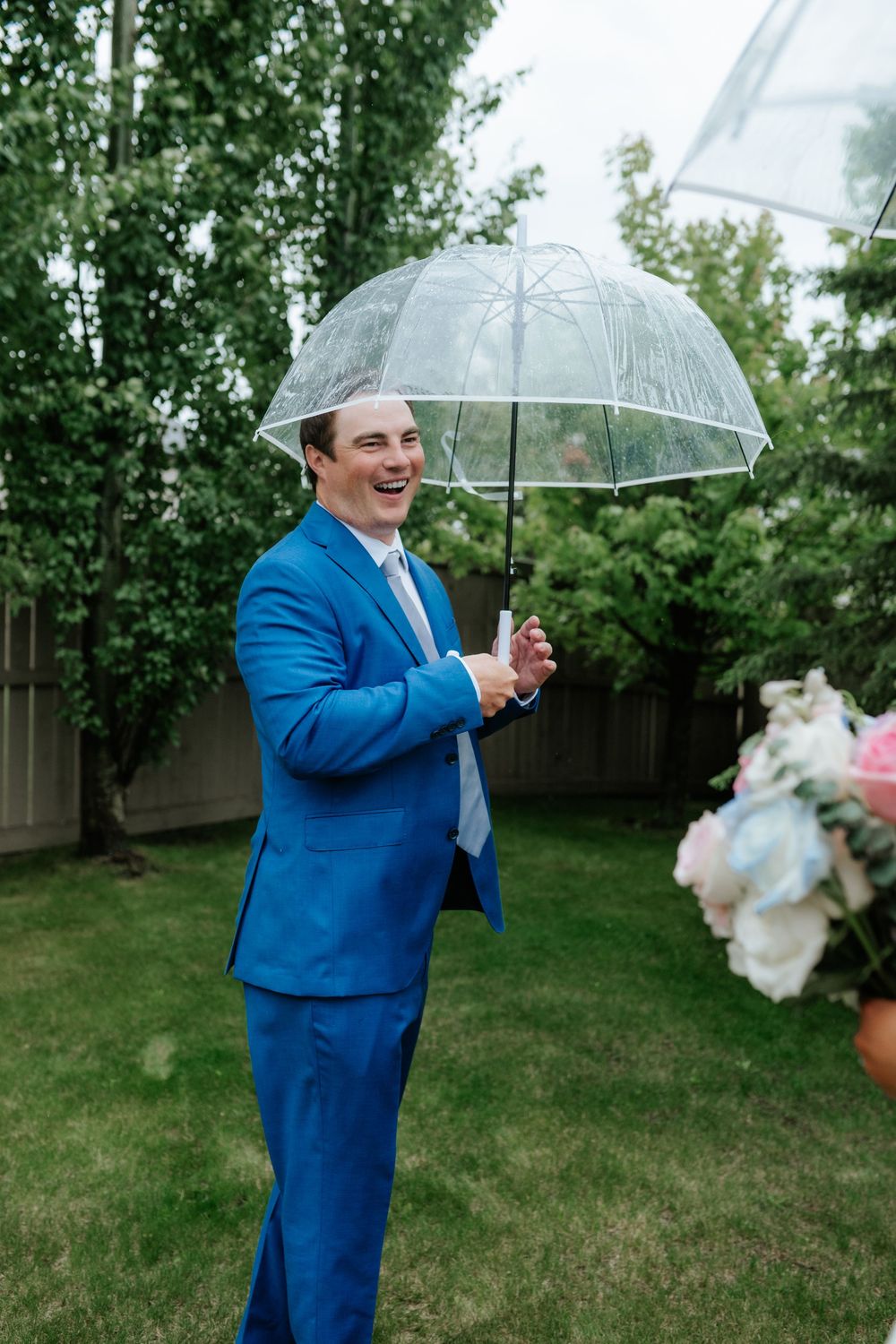 A smiling groom in a blue suit holds a clear umbrella in a green backyard while a bouquet of pastel flowers is visible nearby.