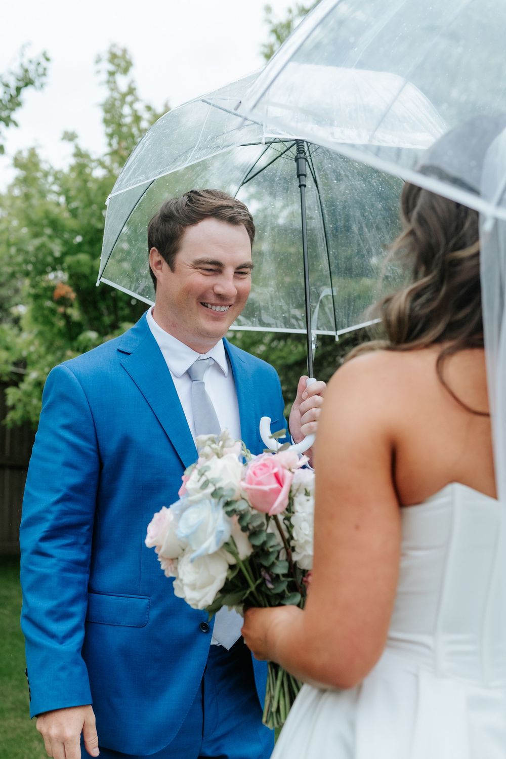 Smiling groom in a blue suit stands under a clear umbrella facing a bride in a white gown holding a pink and white bouquet.