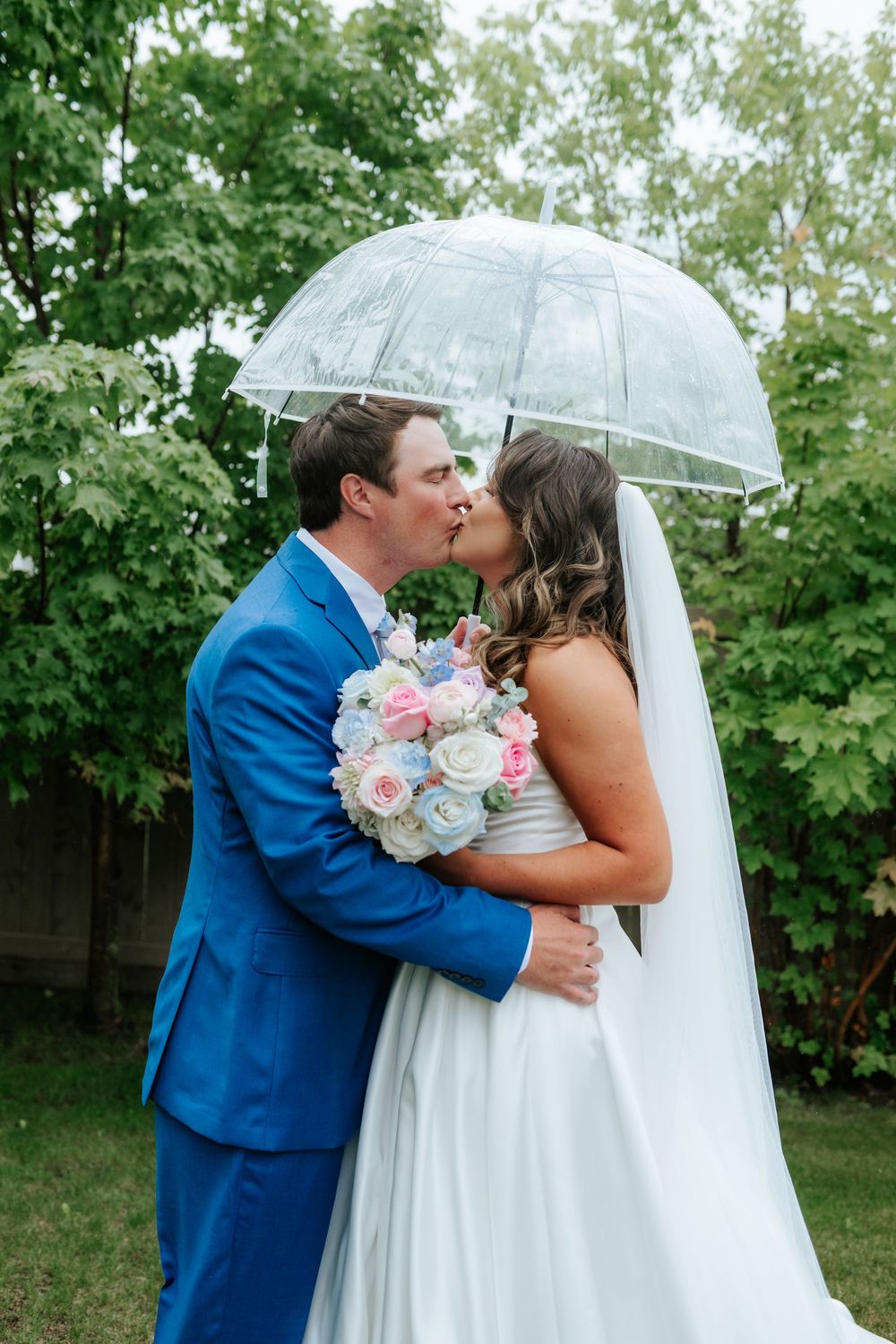 Bride and groom in a blue suit kiss under a clear umbrella, holding a pastel pink and blue bouquet in a leafy green garden.