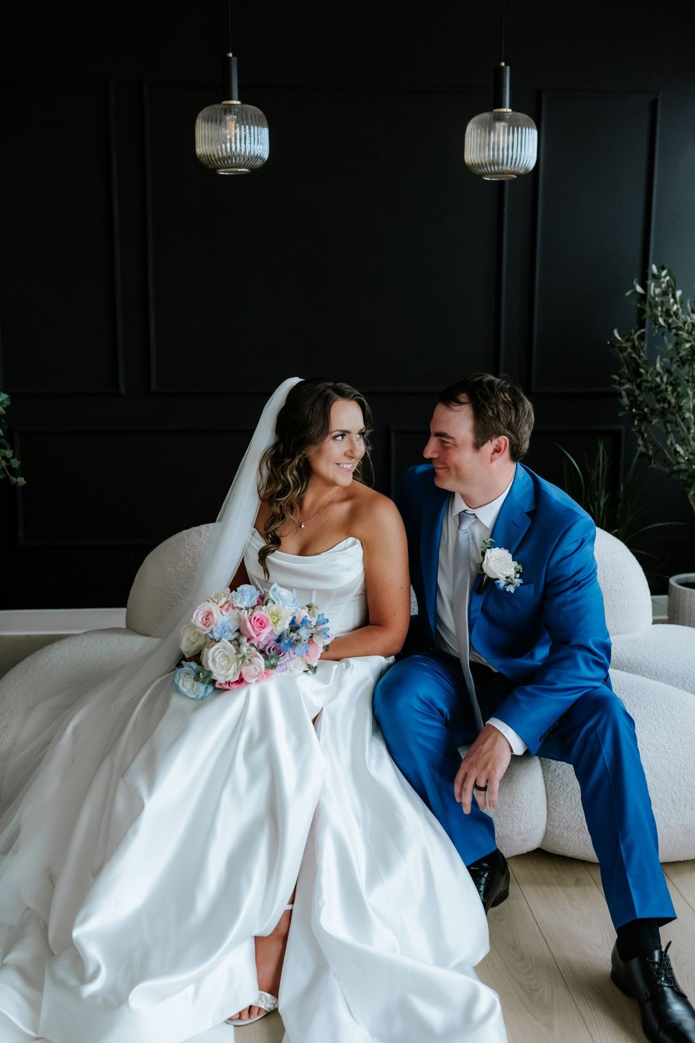 A bride in a white gown holding a pastel bouquet sits beside her groom in a blue suit on a cream sofa against a dark paneled wall.