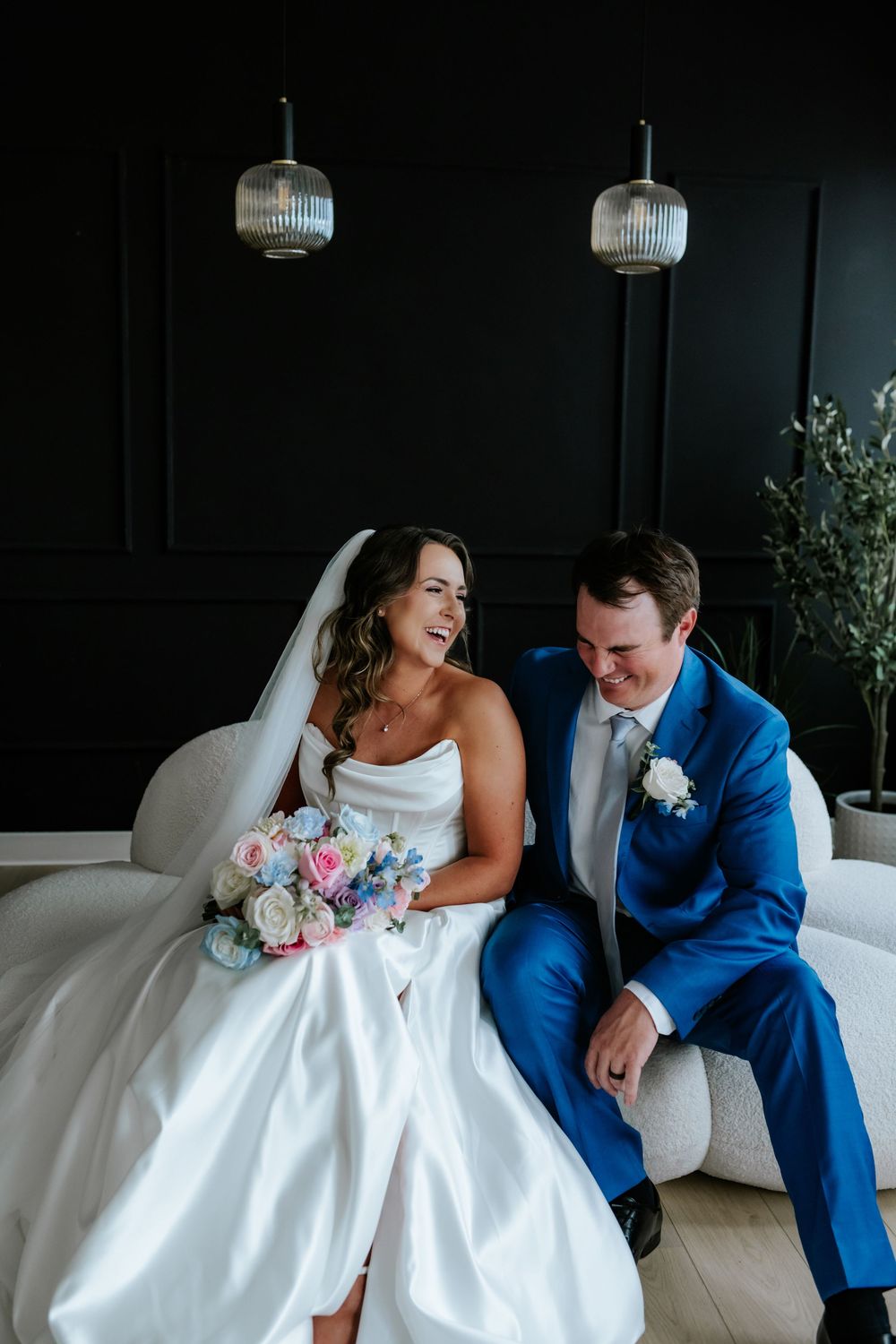 Smiling bride in a satin gown and veil holding a pastel bouquet laughs beside her groom in a blue suit on a white sofa.