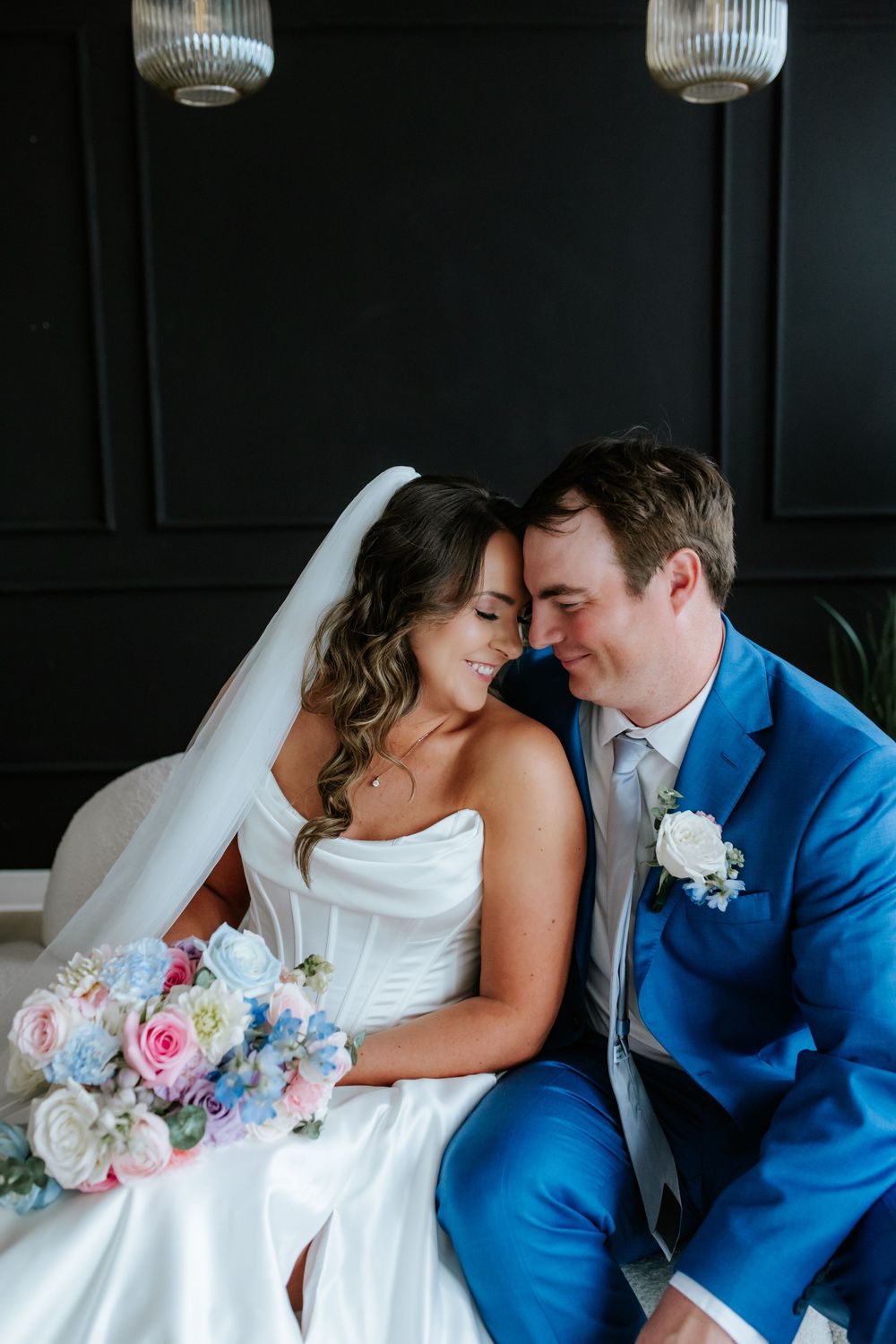 Smiling bride in a white strapless gown and veil holding a pastel bouquet leans foreheads together with a groom in a bright blue suit.